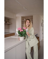 Une femme vêtue d'un costume vert clair se tient dans une cuisine moderne et sourit tout en tenant la plante d'hortensia artificielle rose UV de 35 cm sur le comptoir.