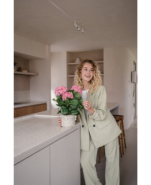 Une femme vêtue d'un costume vert clair se tient dans une cuisine moderne et sourit tout en tenant la plante d'hortensia artificielle rose UV de 35 cm sur le comptoir.