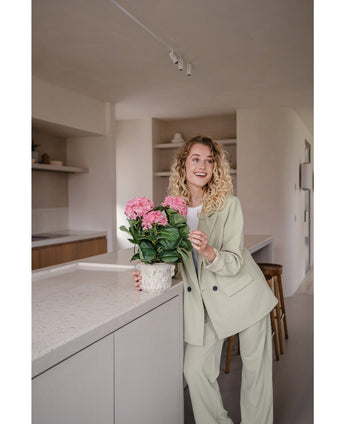 Une femme vêtue d'un costume vert clair se tient dans une cuisine moderne et sourit tout en tenant la plante d'hortensia artificielle rose UV de 35 cm sur le comptoir.