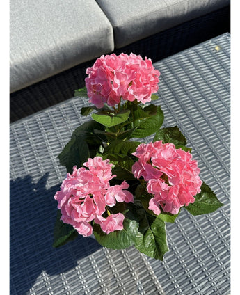 La plante Hortensia Artificielle Rose UV 35 cm, avec trois fleurs d'hortensia roses et des feuilles vertes, se dresse sur une table d'extérieur grise tressée, avec un canapé gris avec des coussins en arrière-plan.
