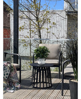 Une terrasse moderne avec deux chaises avec coussins, une table ronde noire avec la plante Laurier Artificiel UV d33 cm comme pièce maîtresse et une couverture à motifs sur une chaise. Des arbres et une clôture en bois apparaissent en arrière-plan sous un ciel dégagé.