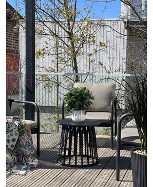 Une terrasse moderne avec deux chaises avec coussins, une table ronde noire avec la plante Laurier Artificiel UV d33 cm comme pièce maîtresse et une couverture à motifs sur une chaise. Des arbres et une clôture en bois apparaissent en arrière-plan sous un ciel dégagé.