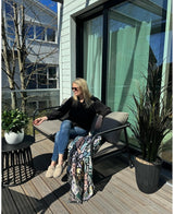 Une femme portant des lunettes de soleil, un haut noir et un jean bleu se détend sur une chaise rembourrée sur un balcon ensoleillé avec un verre à la main. La chambre est équipée de la plante Laurier Artificiel UV d33 cm, d'un décor vivant, d'un plaid coloré et d'une petite table avec des plantes.