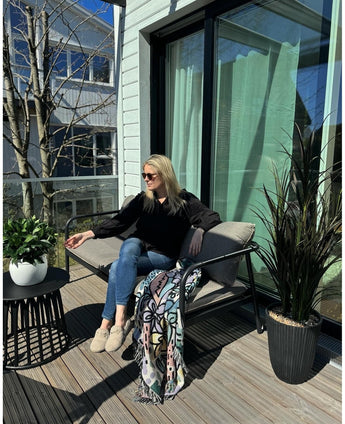 Une femme portant des lunettes de soleil, un haut noir et un jean bleu se détend sur une chaise rembourrée sur un balcon ensoleillé avec un verre à la main. La chambre est équipée de la plante Laurier Artificiel UV d33 cm, d'un décor vivant, d'un plaid coloré et d'une petite table avec des plantes.