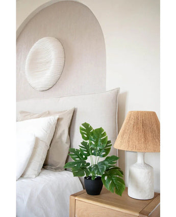 Une chambre moderne aux tons neutres comprend un lit blanc avec des oreillers beiges et blancs, une table de chevet en bois avec une lampe en céramique et un abat-jour tressé, une décoration murale abstraite au-dessus du lit et l'Art Monstera 40 cm dans un pot décoratif noir qui ajoute de la verdure.