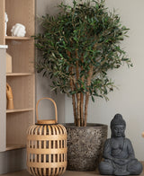 Un olivier artificiel UV 120 cm se dresse à côté d'une statue de Bouddha grise et d'une lanterne en bois au sol, à côté d'une planche de bois clair avec des ornements en forme de main. Cette plante nécessitant peu d’entretien apporte une atmosphère calme et sereine à la pièce.