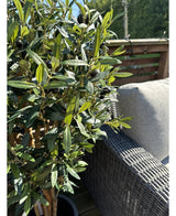 L'Olivier artificiel UV 120 cm aux feuilles vertes et olives noires se dresse à côté d'un canapé d'extérieur en osier gris sur une terrasse ensoleillée avec balustrade en bois.