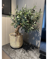 Le support d'olivier artificiel UV 60 cm, avec des feuilles vertes et des olives noires, se trouve dans un panier tressé sur une surface en marbre foncé contre un mur lambrissé clair - facile à entretenir et résistant aux UV pour une beauté durable.