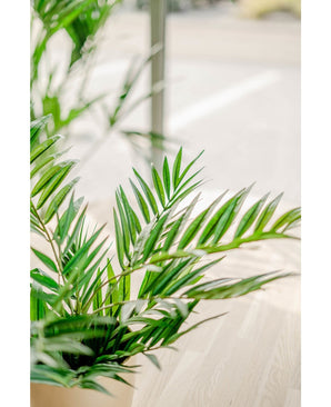 Un gros plan de feuilles de palmier artificiel vert vif UV 60 cm dans un espace intérieur ensoleillé avec un plancher en bois clair et un arrière-plan flou, soulignant la beauté de cette plante artificielle réaliste.