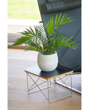 Un palmier artificiel Parlour UV 60 cm se dresse dans un pot sur une petite table d'appoint moderne avec un plateau noir et des pieds en fil métallique, à côté d'une chaise noire sur un parquet en bois clair.