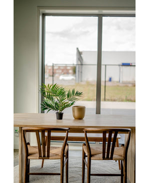 Une table en bois avec deux chaises, un salon de palmiers artificiels UV 60 cm dans un pot et un vase doré sont placés près d'une grande fenêtre donnant sur une zone industrielle avec des bâtiments et une clôture.