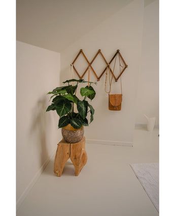 Un coin de pièce minimaliste devient une oasis de verdure avec la plante artificielle Syngonium UV 58 cm sur un tabouret en bois naturel, à côté d'un sac en cuir marron sur une étagère murale géométrique et d'une cruche blanche, le tout sur des murs et un sol blanc cassé.