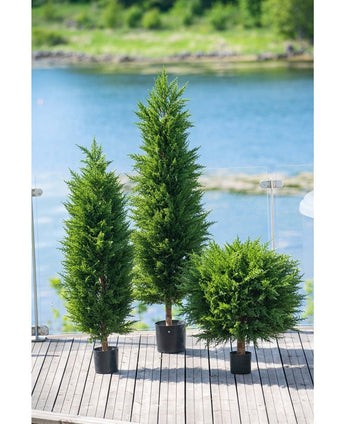 Trois arbres de conifères artificiels de luxe UV de 120 cm ornent une terrasse en bois avec balustrade en verre, surplombant l'eau et la verdure - idéaux pour tout jardin de plantes artificielles grâce à leur protection UV.