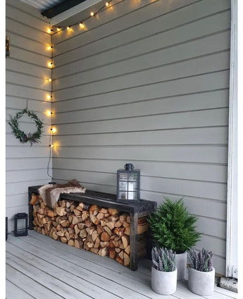 Un coin extérieur confortable comprend un banc fait de bois de chauffage empilé, une lanterne sur le dessus, la plante artificielle Conifer deluxe UV 60 cm, des plantes en pot à proximité, une couronne sur le mur et une guirlande lumineuse au-dessus contre des murs en bois gris clair.