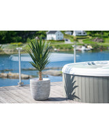 Een Kunstplant Yucca 100 cm UV staat op een houten dek naast een hot tub met glazen reling, met uitzicht op een rustig meer en groene heuvels bezaaid met huizen.