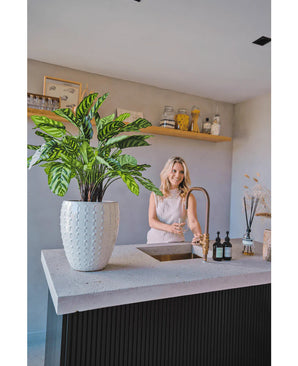 Une femme sourit devant un comptoir de cuisine moderne avec sa main sous le robinet. La Plante Artificielle Calathea Makoyana Deluxe UV 75 cm est au premier plan sur le comptoir. Des étagères ouvertes présentent des pots et des objets décoratifs en arrière-plan.