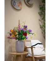 Een Kunstkornoelje Real Touch Lichtgeel vaasje van 74 cm met lichtgele bloemen staat op een houten tafeltje naast een stoel met een wit kussen en een deken. Geweven manden sieren de beige muur en daarachter is een bladplant zichtbaar.