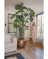 Une femme en tenue beige se tient à côté de la plante artificielle Monstera de 240 cm dans un pot en bronze. Le salon moderne dispose d'un canapé blanc, de grandes fenêtres avec stores et d'un parquet en bois.