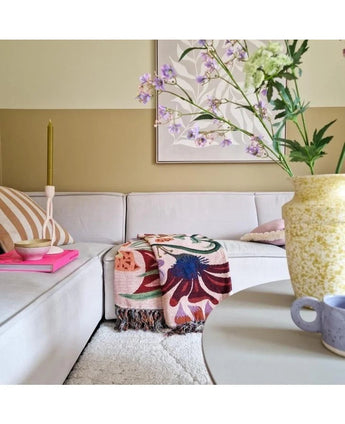 Un salon cosy avec un canapé beige clair, une couverture colorée avec des fleurs et des oreillers à motifs. Sur une table ronde se trouvent un vase jaune avec Kunsttak Daphne Spray 108 cm violet et une petite tasse bleue. Des œuvres d'art sont accrochées au mur aux tons neutres.