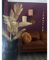 Un salon moderne avec un canapé en velours marron, un coussin rond pliant et le palmier artificiel Areca doré 120 cm dans un vase noir en guise de décor doré. Des œuvres d'art murales géométriques sont accrochées sur un mur violet foncé, avec du parquet et des lattes verticales en vue.