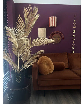 Un salon moderne avec un canapé en velours marron, un coussin rond pliant et le palmier artificiel Areca doré 120 cm dans un vase noir en guise de décor doré. Des œuvres d'art murales géométriques sont accrochées sur un mur violet foncé, avec du parquet et des lattes verticales en vue.