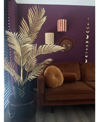 Un salon moderne avec un canapé en velours marron, un coussin rond pliant et le palmier artificiel Areca doré 120 cm dans un vase noir en guise de décor doré. Des œuvres d'art murales géométriques sont accrochées sur un mur violet foncé, avec du parquet et des lattes verticales en vue.