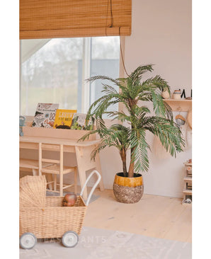 In een gezellige, lichte kamer staan de Kunstpalm Cycas Classic 135 cm in een stijlvolle plantenbak, een rieten speelgoedkar, houten meubels en kinderboeken bij een raam met een bamboe rolgordijn.