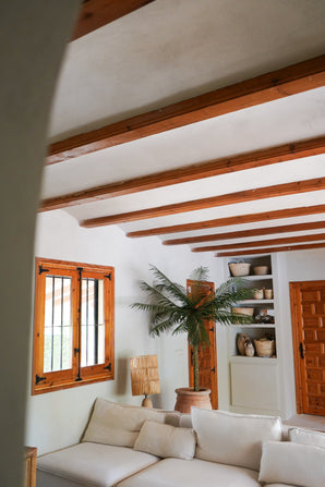 Un salon confortable avec des murs blancs, des poutres en bois, un canapé sectionnel blanc, le palmier artificiel Phoenix De Luxe 210 cm, une fenêtre et une porte en bois, des étagères intégrées avec des paniers et des poteries et de la lumière naturelle entrant par la fenêtre.