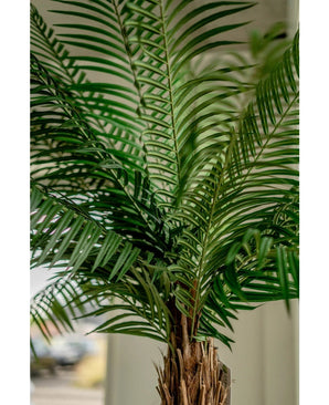Gros plan du palmier artificiel Phoenix De Luxe 210 cm, avec de longues feuilles vertes étroites et un tronc marron texturé, sur un fond légèrement flou.