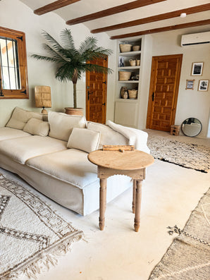 Un salon cosy avec un canapé blanc, une table d'appoint ronde en bois, un palmier artificiel Phoenix De Luxe 210 cm, des paniers tressés sur des étagères, des portes en bois, un miroir rond et des tapis aux tons neutres sur un sol clair.