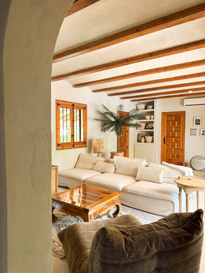 Un salon confortable comprend un grand canapé de couleur crème, une table basse en bois, un palmier artificiel Phoenix De Luxe de 210 cm dans un pot, des poutres apparentes au plafond, des étagères intégrées avec des poteries, une lumière naturelle des fenêtres et un décor chaleureux et rustique.