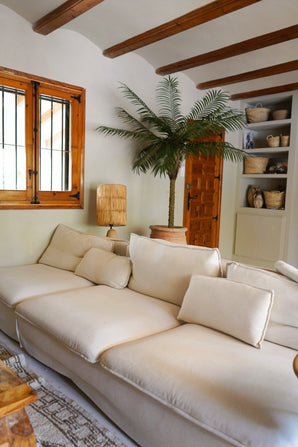 Un salon cosy avec un grand canapé couleur crème, le Palmier Artificiel Phoenix De Luxe 210 cm dans le coin, une lampe en bois, des étagères intégrées avec paniers, des poutres en bois au plafond et une fenêtre avec des cadres en bois.
