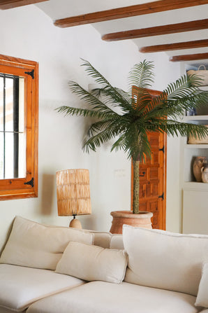 Un salon cosy avec un canapé blanc, une lampe de table en rotin, le Palmier Artificiel Phoenix De Luxe 210 cm dans un pot, des poutres en bois au plafond, une fenêtre avec des cadres en bois et une porte en bois en arrière-plan.