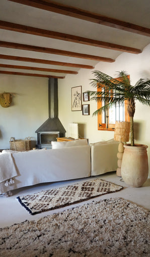 Un salon confortable dispose d'un canapé blanc, d'un tapis à motifs, d'un poêle à bois, d'œuvres d'art encadrées et de poutres en bois. La lumière du soleil pénètre depuis une fenêtre à côté du luxuriant palmier artificiel Phoenix De Luxe de 210 cm, donnant vie à l'espace.