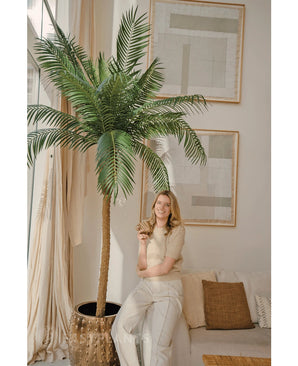 Une femme aux cheveux blonds portant un haut beige et un pantalon clair sourit en s'appuyant contre le palmier artificiel Phoenix De Luxe 210 cm dans un salon lumineux et moderne avec une décoration neutre et des œuvres d'art abstraites sur les murs.