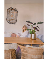 Une femme en blouse blanche est assise à une table à manger en bois avec des chaises en osier. La pièce confortable et doucement éclairée est dotée de la plante artificielle Alocasia UV 67 cm et d'une lampe suspendue texturée comme décor.