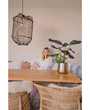 Une femme en blouse blanche est assise à une table à manger en bois avec des chaises en osier. La pièce confortable et doucement éclairée est dotée de la plante artificielle Alocasia UV 67 cm et d'une lampe suspendue texturée comme décor.