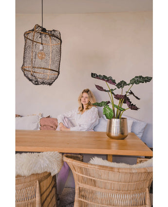 Une femme en blouse blanche est assise à une table à manger en bois avec des chaises en osier. La pièce confortable et doucement éclairée est dotée de la plante artificielle Alocasia UV 67 cm et d'une lampe suspendue texturée comme décor.