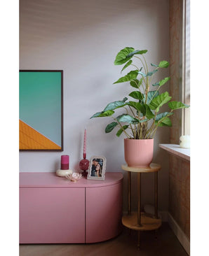 Un coin salon moderne avec un buffet rose, des bougies, une décoration murale géométrique, une photo encadrée et la plante artificielle Feuille de pommier de 90 cm - une grande plante à feuillage dans un pot rose sur un petit support en bois près de la fenêtre.