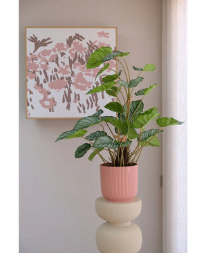 La Feuille de Pommier Artificielle 90 cm, une plante en pot à feuilles vertes dans un pot rose nervuré, se dresse sur un socle beige devant un mur avec un art floral rose abstrait et un rideau blanc translucide à droite.