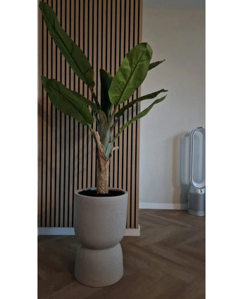 De Kunstplant Bananenboom 150 cm met grote groene bladeren staat op een houten vloer voor een houten wand met latten. Rechts staat een moderne bladloze ventilator in de kamer met neutrale muren en vloer.