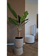 De Kunstplant Bananenboom 150 cm, met zijn weelderige groene bladeren, staat op een houten vloer bij een geribbelde wand met houten panelen. Vlakbij staan een witte ronde voetenbank en een ventilator zonder bladen op de achtergrond.