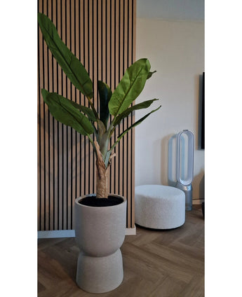 De Kunstplant Bananenboom 150 cm, met zijn weelderige groene bladeren, staat op een houten vloer bij een geribbelde wand met houten panelen. Vlakbij staan een witte ronde voetenbank en een ventilator zonder bladen op de achtergrond.