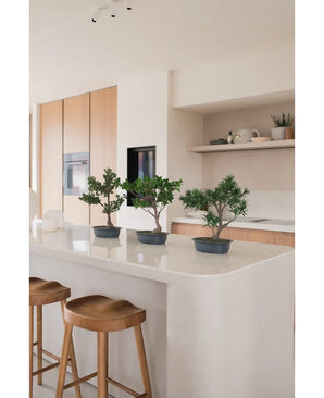 Une plante artificielle Bonsai Ficus de 40 cm se dresse sur un îlot de cuisine blanc avec deux tabourets en bois, entourés d'armoires en bois clair, de plans de travail et d'étagères blancs au décor minimaliste.