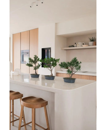 Une plante artificielle Bonsai Juniper de 40 cm se dresse sur un îlot de cuisine blanc avec deux tabourets en bois, entourés d'armoires en bois clair, de plans de travail blancs et d'un décor minimaliste.
