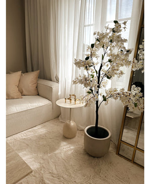 Un coin salon cosy avec un canapé blanc, des coussins beiges, une petite table d'appoint ronde avec des bougeoirs dorés, des rideaux transparents et le Bougainvillier Artificiel 120 cm blanc pour une touche élégante.
