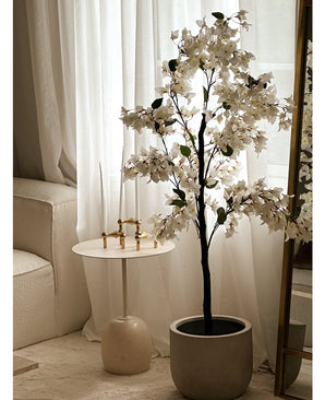 Une plante de bougainvilliers artificiels mesurant 120 cm se trouve près d'un miroir bordé d'or, à côté d'une petite table d'appoint ronde blanche avec des chandeliers dorés, de hautes bougies blanches, des rideaux blancs transparents et un canapé de couleur crème dans une pièce lumineuse.