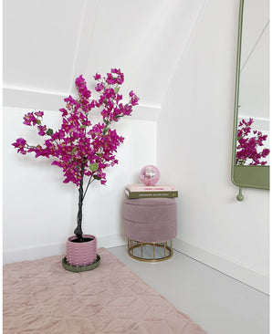 Een moderne, minimale kamerhoek met een Kunstplant Bougainvillea 125 cm Paars in een pot, een rond roze voetenbankje met gouden pootjes, gestapelde boeken, een roze kristallen bol, een wandspiegel en een zachtroze vloerkleed op een lichte vloer.
