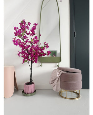 Een modern interieur met de Kunstplant Bougainvillea 125 cm Paars in een decoratieve pot, een hoge spiegel met groene lijst, een roze voetenbank met gouden voet en roze deken, en een donkere deur tegen een kraakheldere witte muur.