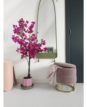 Een modern interieur met de Kunstplant Bougainvillea 125 cm Paars in een decoratieve pot, een hoge spiegel met groene lijst, een roze voetenbank met gouden voet en roze deken, en een donkere deur tegen een kraakheldere witte muur.